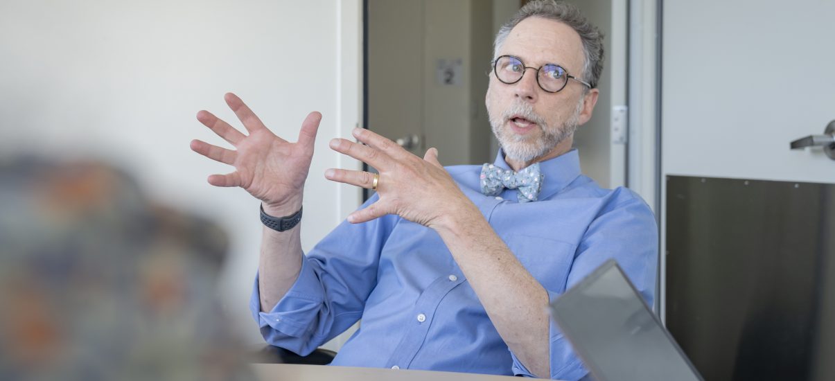 A man in a shirt and bow tie gestures expressively while speaking during a meeting