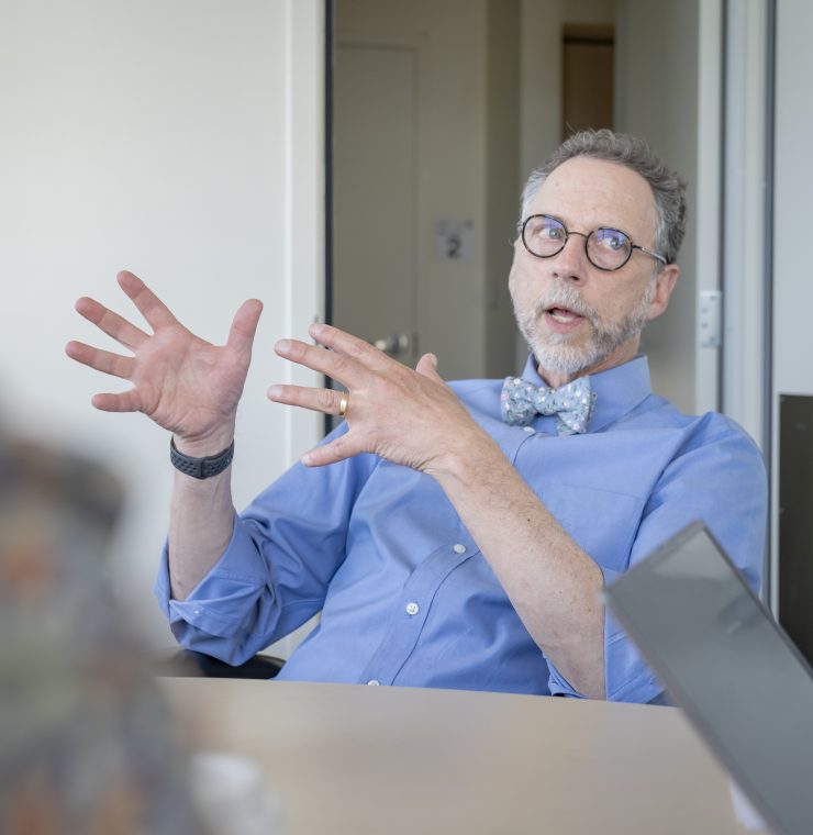 A man in a shirt and bow tie gestures expressively while speaking during a meeting