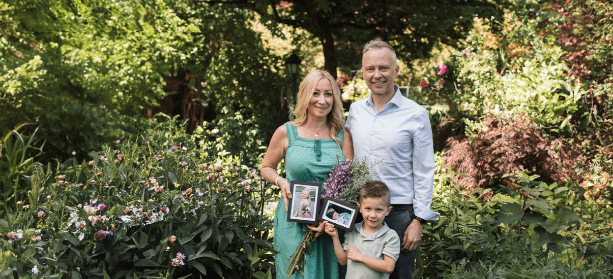 A family of three stands in a lush garden surrounded by blooming flowers. The woman in a green dress holds a bouquet and a framed photo of a young girl. A young boy beside her holds another framed photo of a baby, while a man in a light blue shirt stands next to them smiling. The scene evokes remembrance and love in a peaceful, vibrant setting.