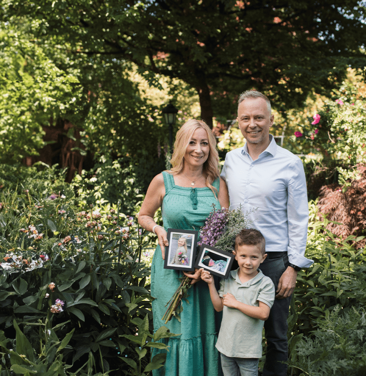 A family of three stands in a lush garden surrounded by blooming flowers. The woman in a green dress holds a bouquet and a framed photo of a young girl. A young boy beside her holds another framed photo of a baby, while a man in a light blue shirt stands next to them smiling. The scene evokes remembrance and love in a peaceful, vibrant setting.