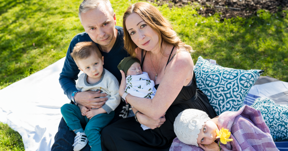 A family sits closely together on a blanket in a sunlit garden. A woman cradles a sleeping baby wrapped in a white blanket with green leaves, while a man holds a toddler on his lap. Beside them, a young girl in a white knit hat lies on a pillow, holding a yellow daffodil in her mouth. The mood is tender and serene, filled with love and gentle connection.