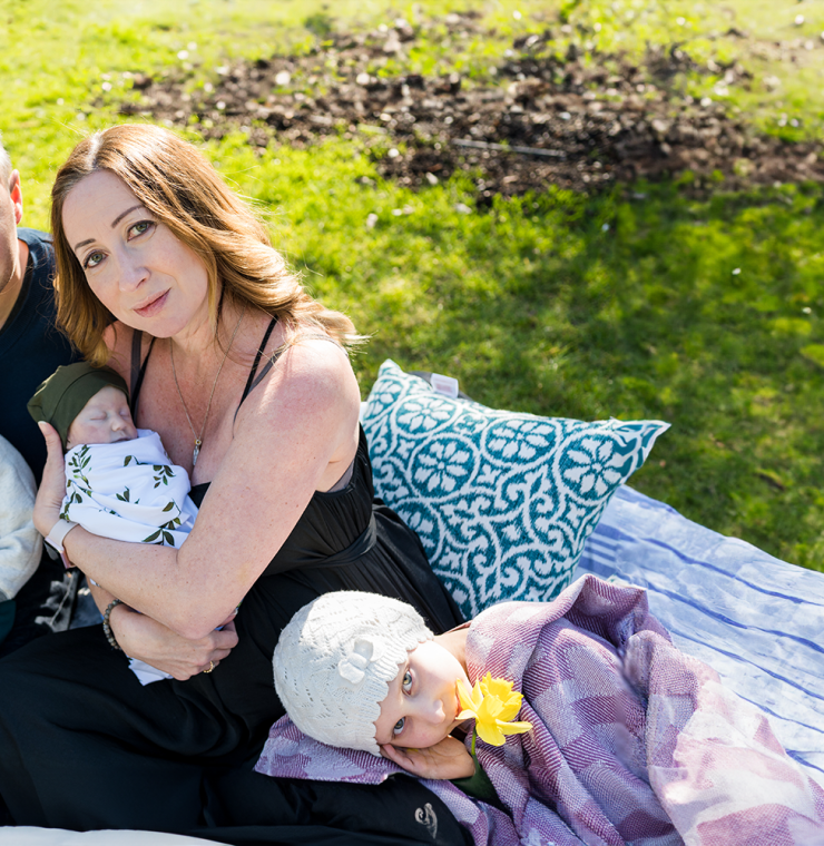 A family sits closely together on a blanket in a sunlit garden. A woman cradles a sleeping baby wrapped in a white blanket with green leaves, while a man holds a toddler on his lap. Beside them, a young girl in a white knit hat lies on a pillow, holding a yellow daffodil in her mouth. The mood is tender and serene, filled with love and gentle connection.