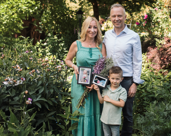 A family of three stands in a lush garden surrounded by blooming flowers. The woman in a green dress holds a bouquet and a framed photo of a young girl. A young boy beside her holds another framed photo of a baby, while a man in a light blue shirt stands next to them smiling. The scene evokes remembrance and love in a peaceful, vibrant setting.