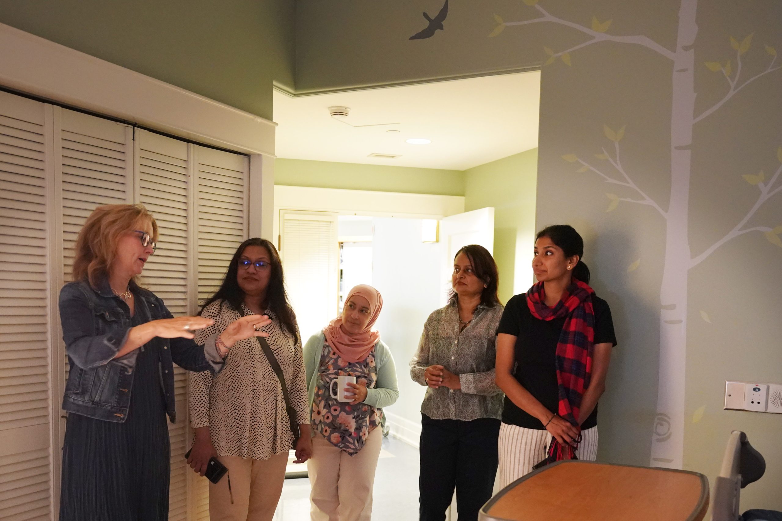 Five women stand together in a softly lit room with green walls and a tree mural. The woman on the far left gestures while speaking, wearing a denim jacket over a dark dress. The other four women listen attentively, with one holding a mug and others standing with hands clasped, dressed in patterned tops and light-colored pants.