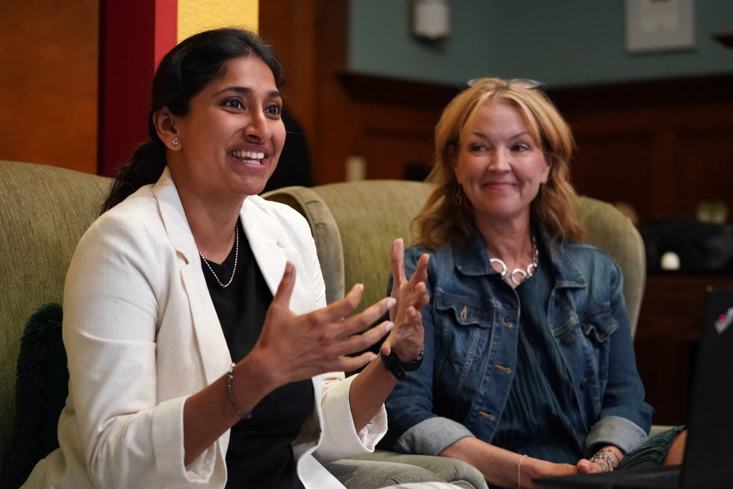 A woman in a white blazer gestures expressively with both hands while speaking, smiling warmly. Next to her, another woman with blonde hair in a denim jacket sits back, watching her with an encouraging smile.