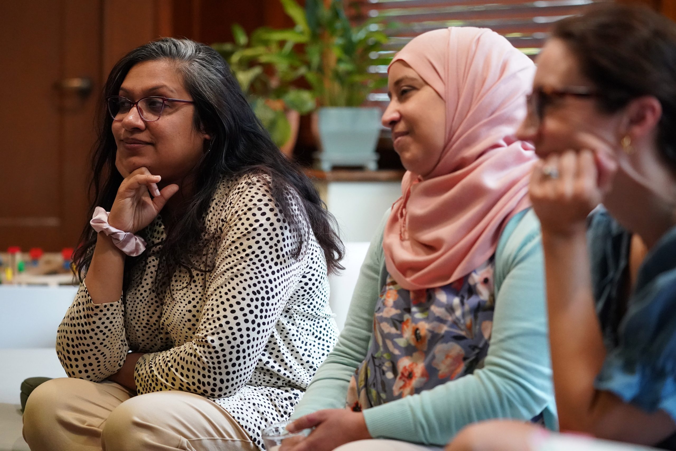 Three women sit closely together, attentively listening. The woman on the left has long dark hair, glasses, and wears a polka-dotted shirt with her chin resting on her hand. The woman in the middle wears a light pink hijab and floral blouse and has a soft smile. The woman on the right, slightly blurred, wears glasses and rests her hand on her cheek, near her mouth.